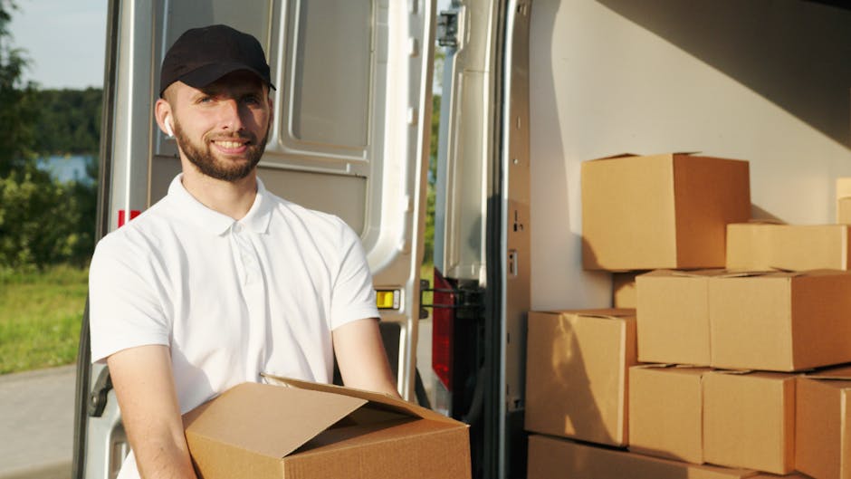 A professional removal technician, wearing a white polo shirt and black cap, stands outdoors beside a moving van with its back doors open. Inside the van, several cardboard boxes of various sizes are