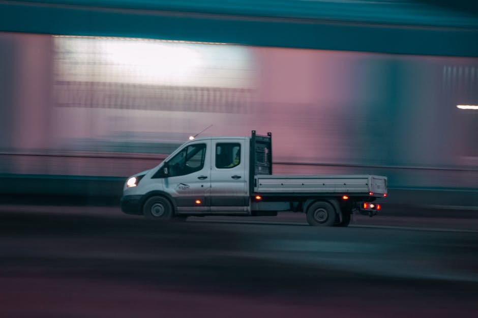 A light grey commercial van is parked on a street in front of a building with arched windows and a sign reading 'M' for Maison Magnum. The van's cargo area is open, revealing a partially extended meta