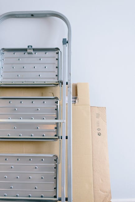 A silver, multi-tiered hand truck or trolley is positioned vertically against a plain white wall inside a home or storage area, with several cardboard boxes of various sizes stacked behind it. The han
