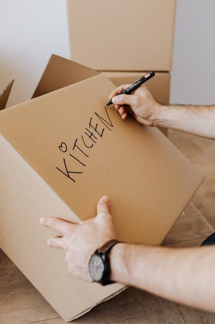 A silver, multi-tiered hand truck or trolley is positioned vertically against a plain white wall inside a home or storage area, with several cardboard boxes of various sizes stacked behind it. The han